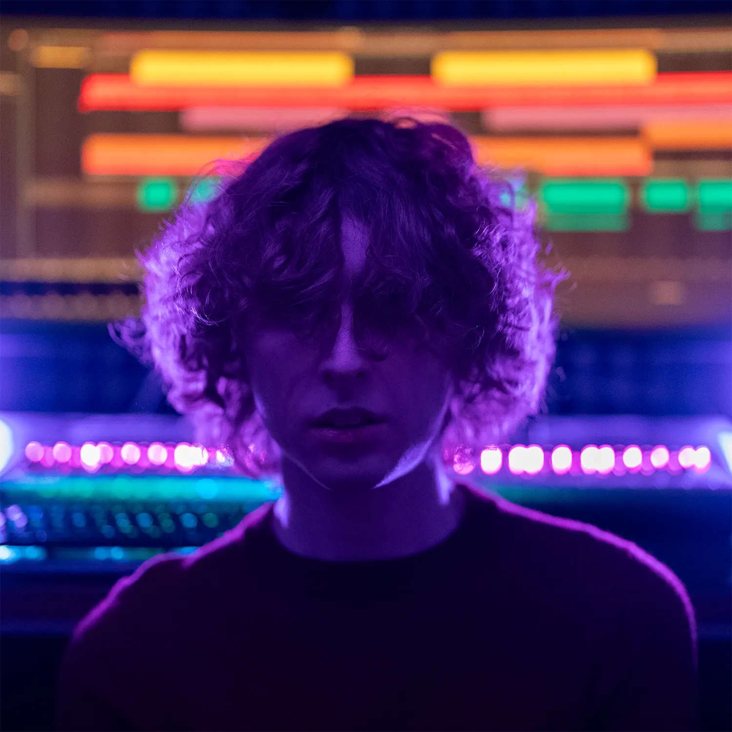 An image of Misha with his curly hair partially covering his face, illuminated by purple and pink neon lights, sitting in front of a blurred background of colorful LED lights and a colorful computer monitor.